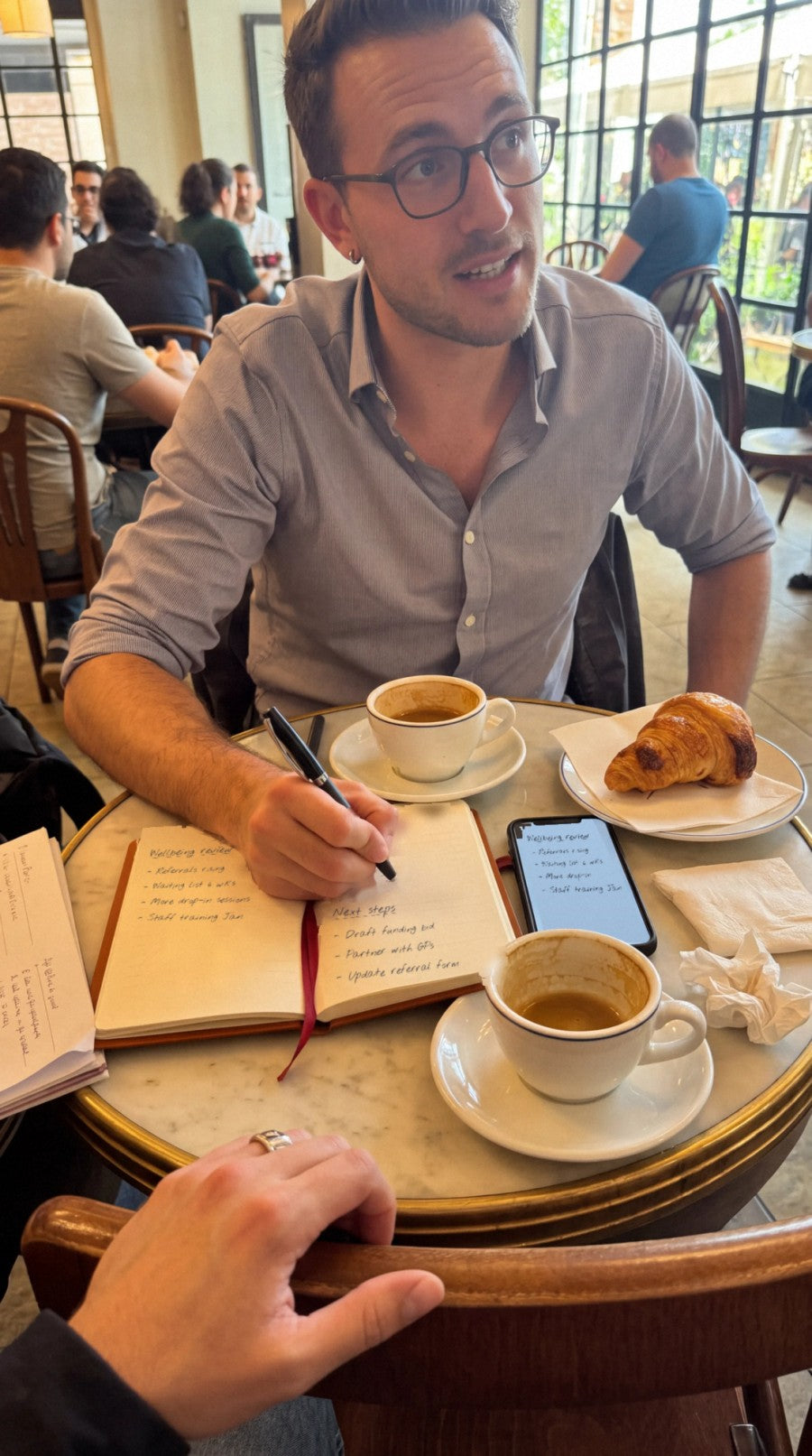 A wellbeing service review over coffee: notes in NoteFlow synced to a phone on the table