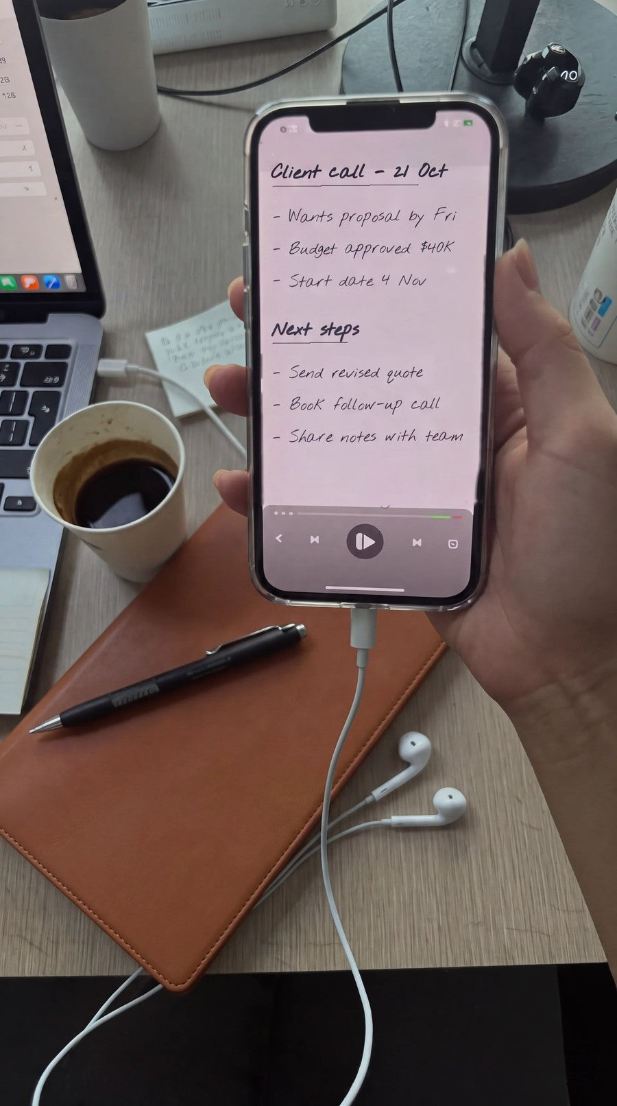 Replaying client call notes on a phone at a desk, with the NoteFlow notebook, pen and earphones